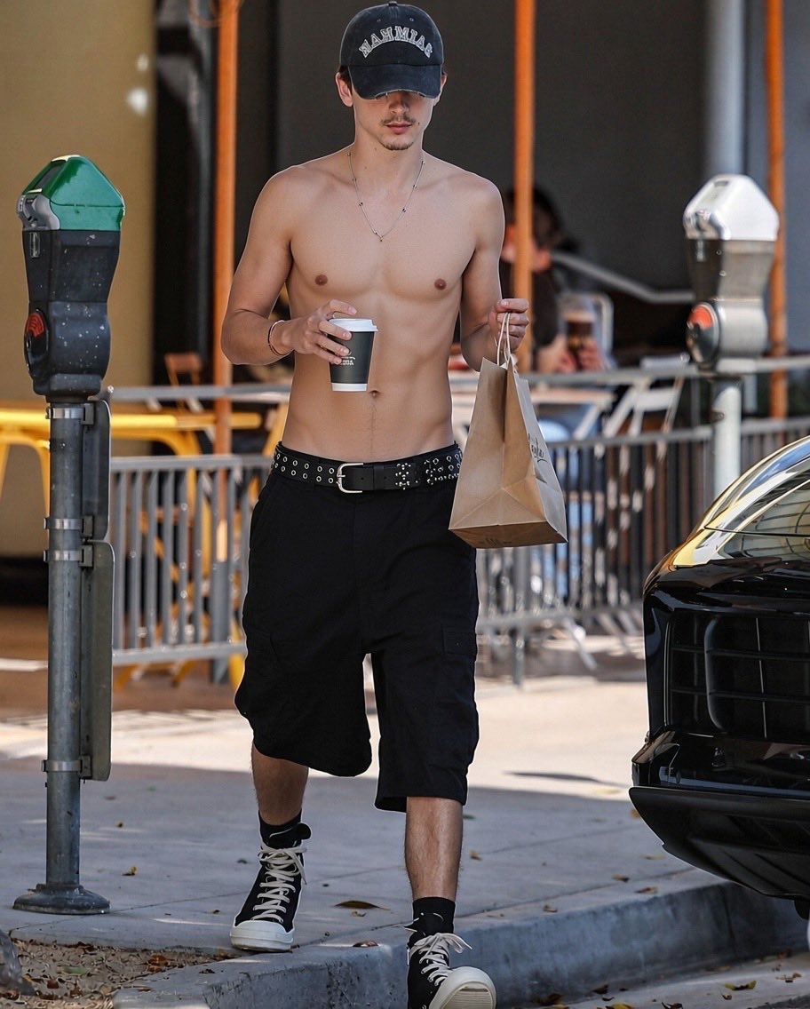 A shirtless man resembling Timothée Chalamet walking on a city sidewalk holding a coffee cup and a paper bag.