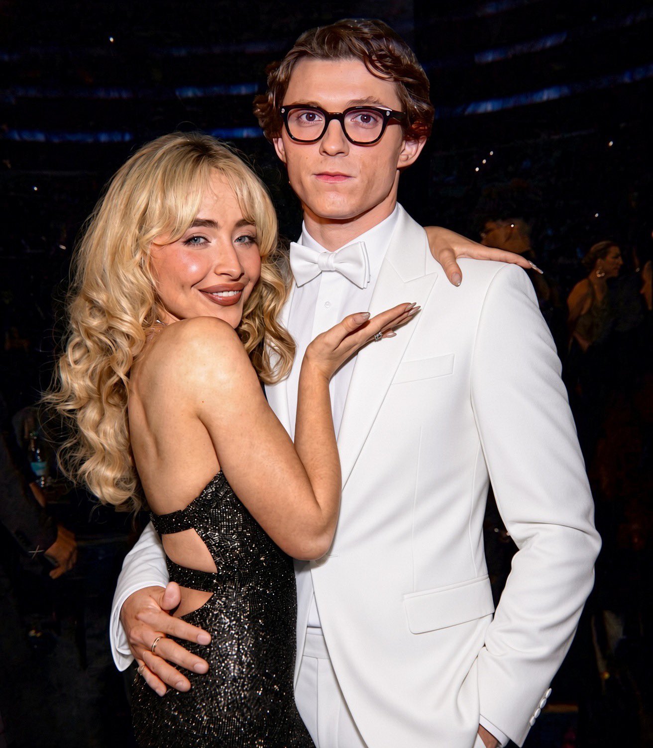 A woman resembling Sabrina Carpenter embracing a man resembling Tom Holland in a white tuxedo at a formal event.