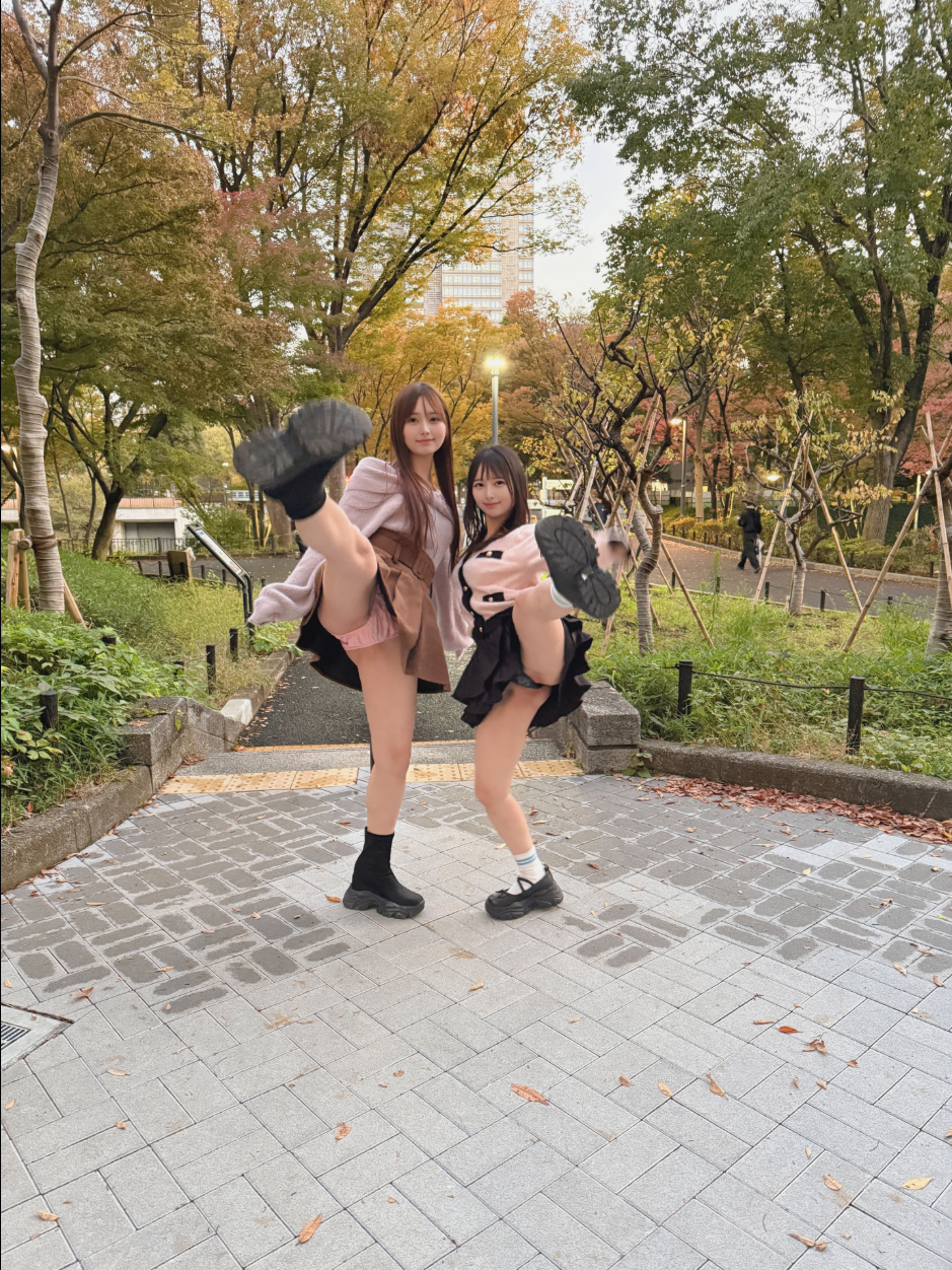 Two young women in skirts kicking their legs toward the camera, soles of their shoes visible, on a paved park walkway.