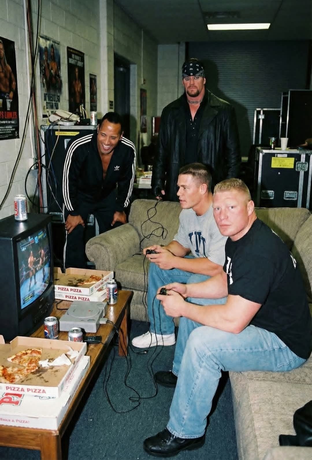 Four men resembling The Undertaker, Dwayne Johnson, John Cena, and Brock Lesnar playing video games in a backstage lounge.