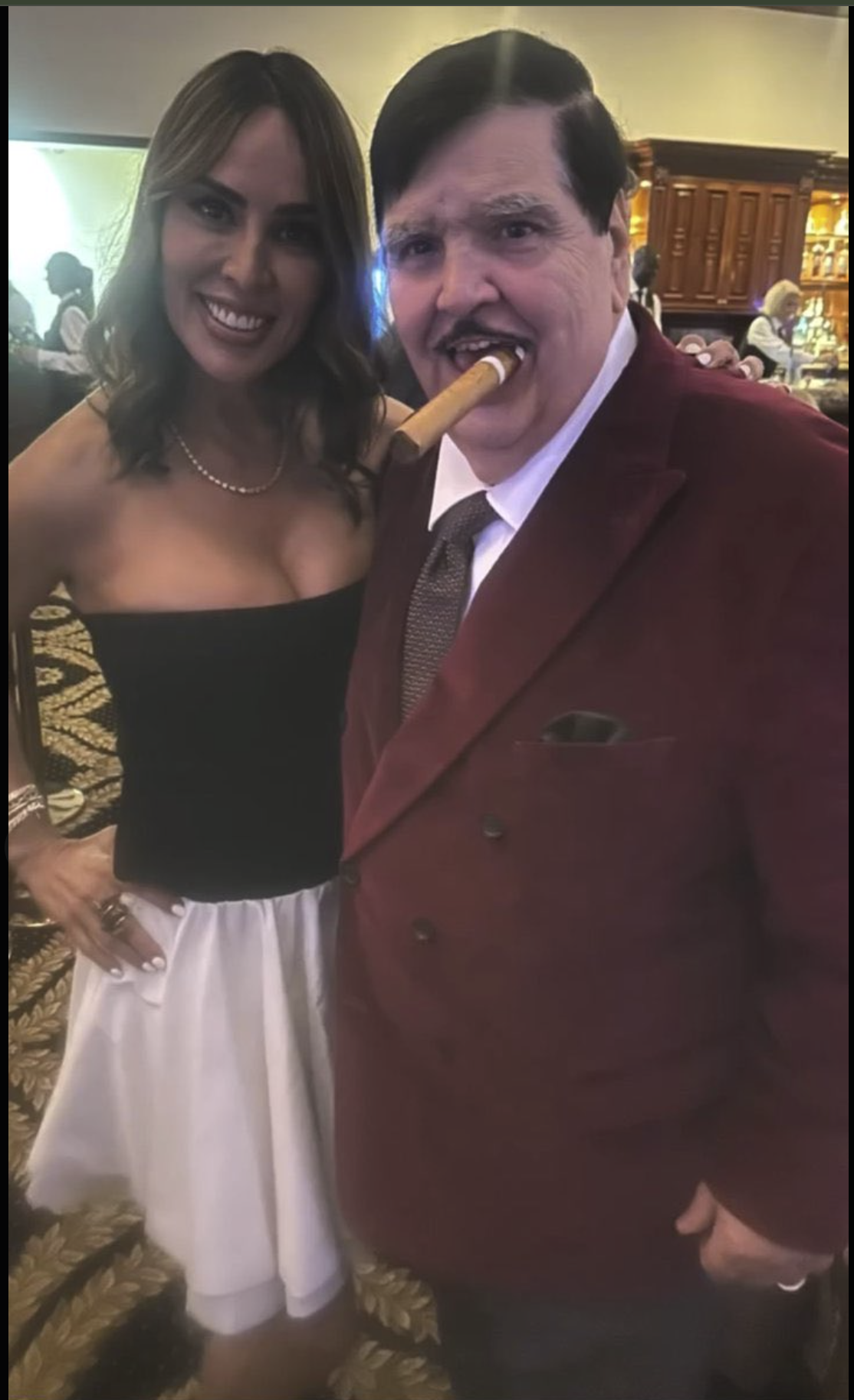 A woman in a black strapless top and a man in a burgundy suit holding a cigar in his mouth, posing together at an indoor event.
