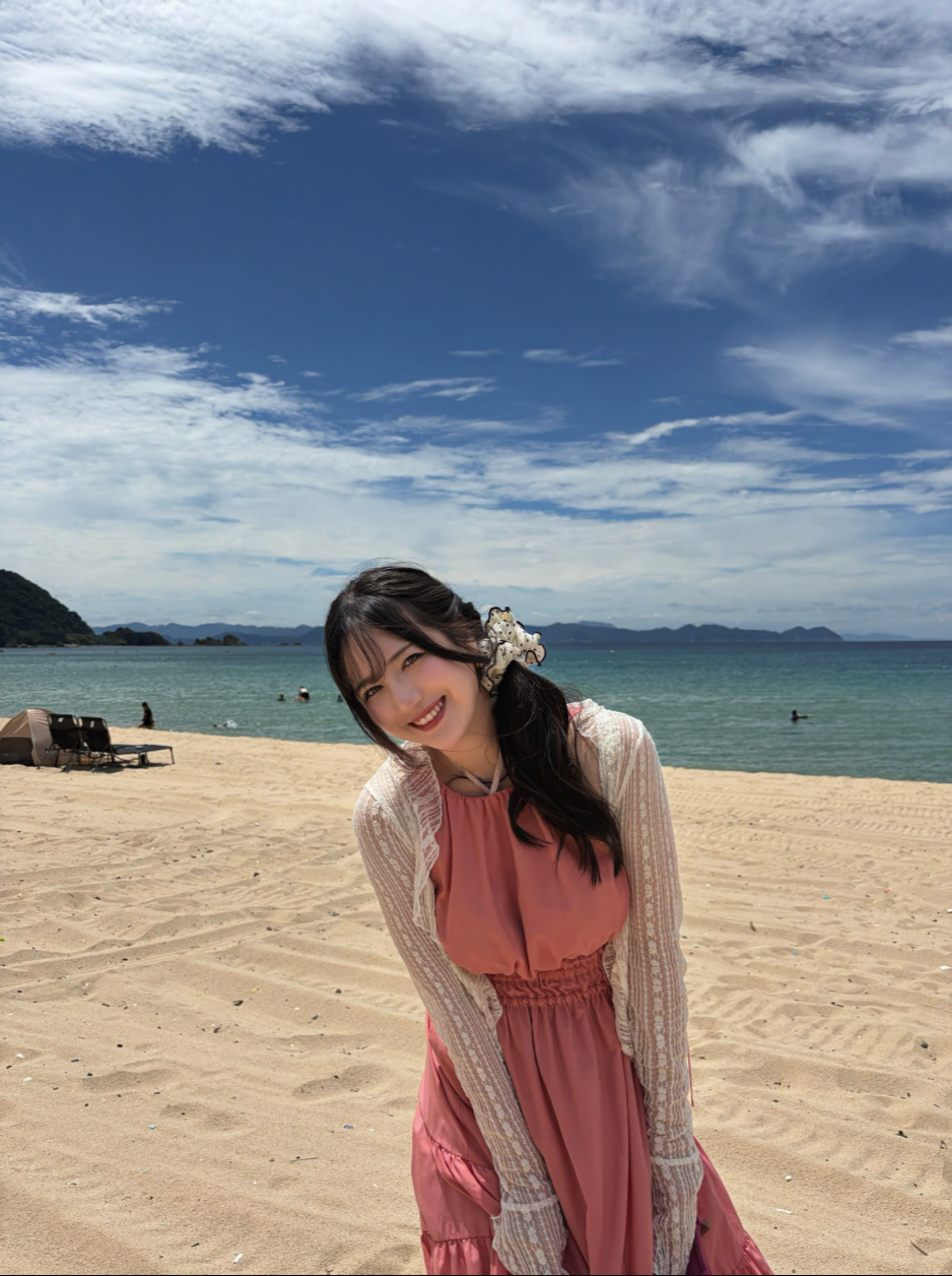 A woman resembling Zhao Jiamin smiling on a sandy beach, wearing a coral dress and a cream lace cardigan.