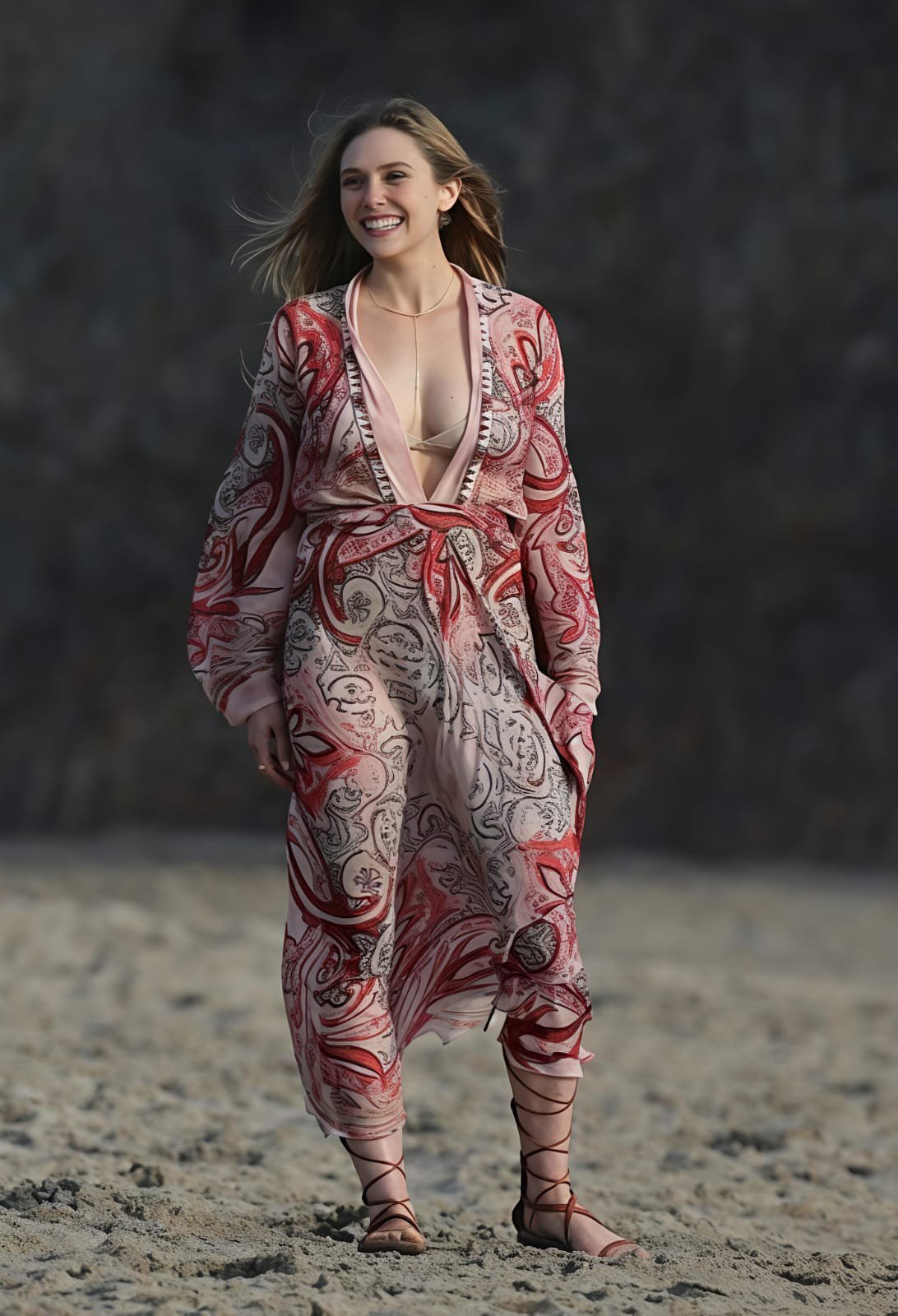 A woman resembling Elizabeth Olsen standing on sand in a flowing red and cream paisley dress.